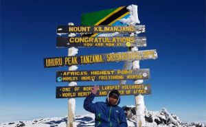 Ein erfolgreicher Bergsteiger sitzt vor dem Schild auf dem Gipfel des Kilimandscharo